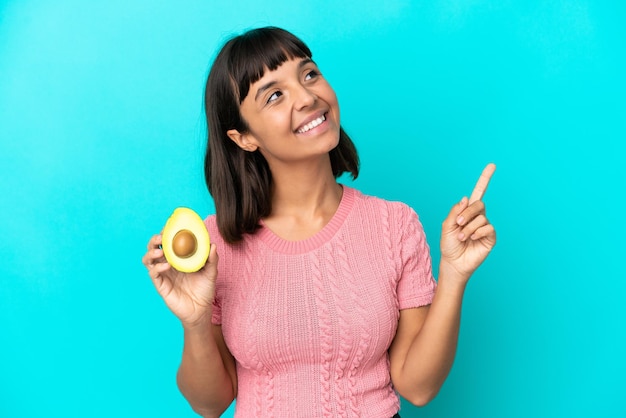 Giovane donna di razza mista in possesso di un avocado isolato su sfondo blu che indica una grande idea