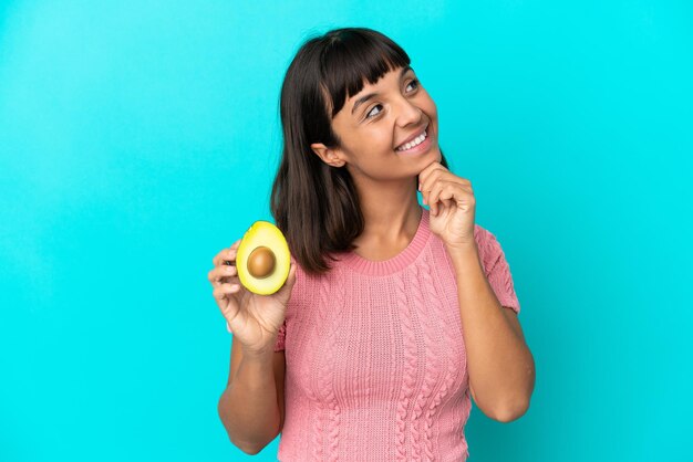 Giovane donna di razza mista in possesso di un avocado isolato su sfondo blu e alzando lo sguardo