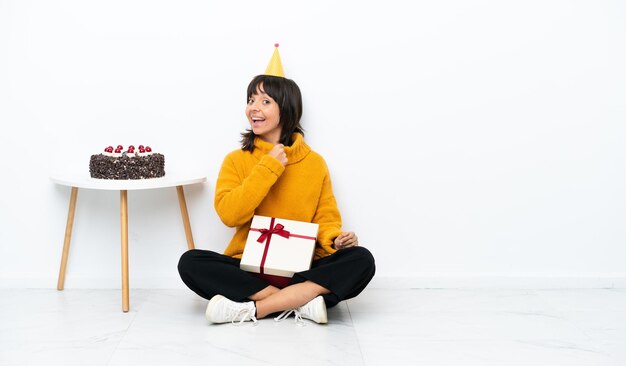 Giovane donna di razza mista in possesso di un regalo seduto sul pavimento isolato su sfondo bianco che celebra una vittoria