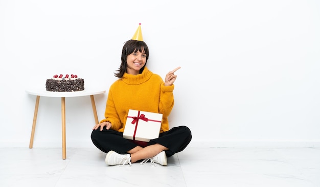 Giovane donna di razza mista in possesso di un regalo seduto sul pavimento isolato su sfondo bianco che punta il dito di lato