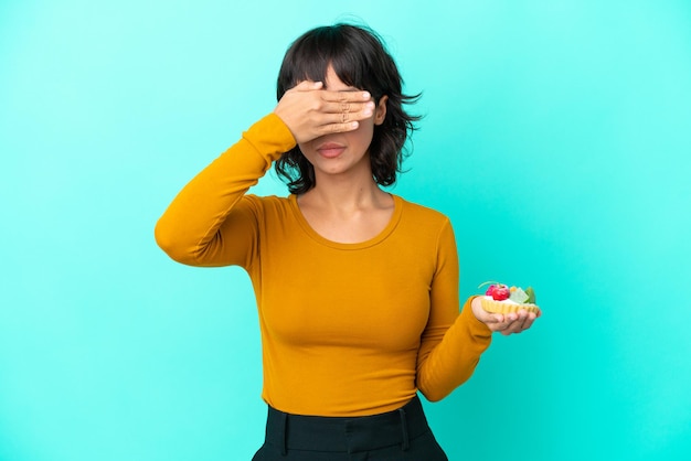 Giovane donna di razza mista in possesso di un tortino isolato su sfondo blu che copre gli occhi con le mani. Non voglio vedere qualcosa