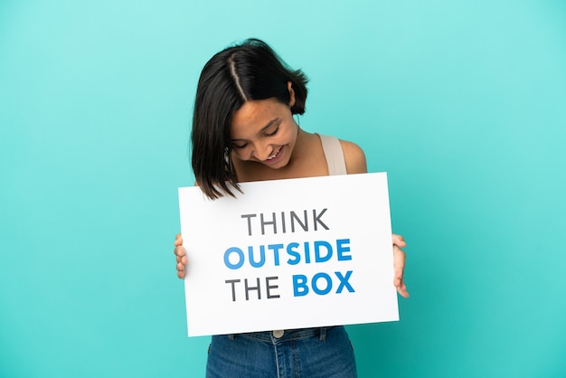 Giovane donna di razza mista isolata su sfondo blu, tenendo un cartello con il testo Think Outside The Box