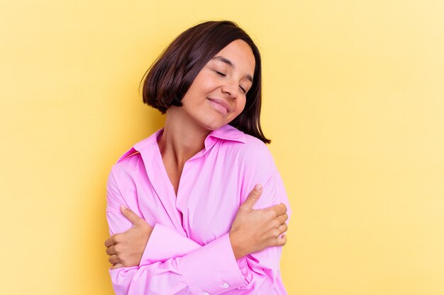 Giovane donna di razza mista isolata su sfondo giallo abbracci, sorridendo spensierata e felice.