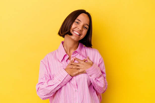 Giovane donna di razza mista isolata su sfondo giallo che ride tenendo le mani sul cuore, concetto di felicità.