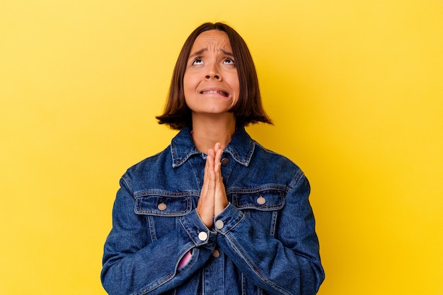 Giovane donna di razza mista isolata su sfondo giallo tenendosi per mano in preghiera vicino alla bocca, si sente sicura.