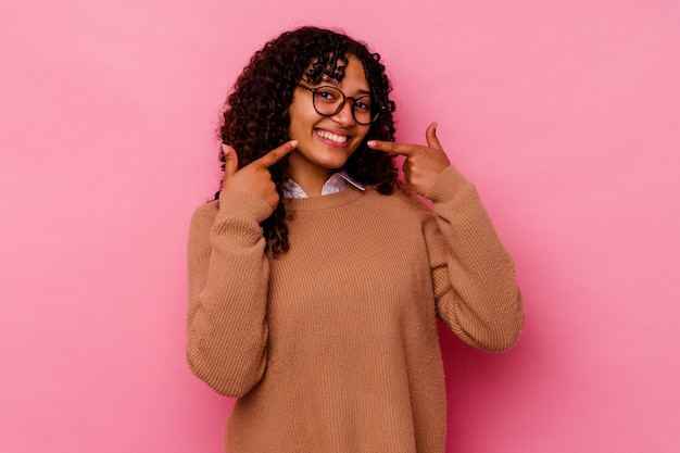 Giovane donna di razza mista isolata su sorrisi rosa, puntando il dito alla bocca.