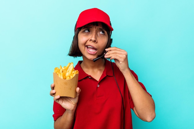 Giovane donna di razza mista ristorante fast food lavoratore tenendo patatine isolate su sfondo blu cercando di ascoltare un pettegolezzo.