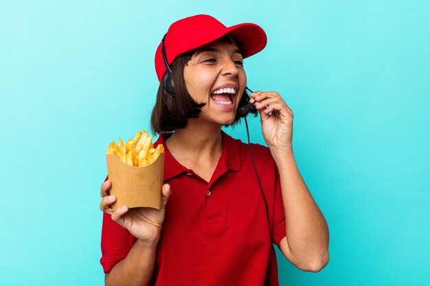 Giovane donna di razza mista ristorante fast food lavoratore tenendo patatine isolate su sfondo blu gridando e tenendo il palmo vicino alla bocca aperta.