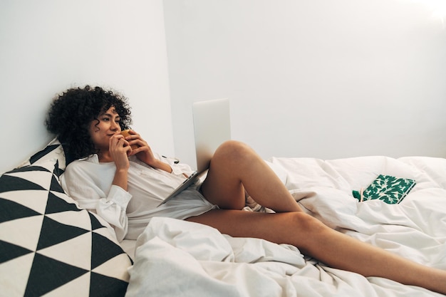 Giovane donna di razza mista sdraiata sul letto usando il computer portatile e bevendo caffè. Copia spazio. Stile di vita e concetto di tecnologia.
