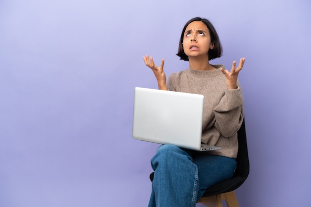 Giovane donna di razza mista seduta su una sedia con laptop isolato su sfondo viola ha sottolineato sopraffatto