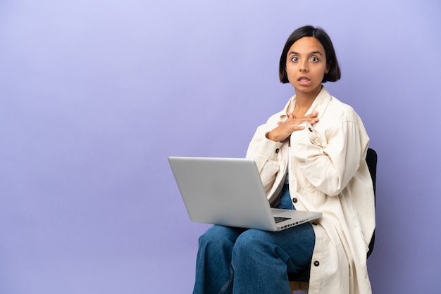 Giovane donna di razza mista seduta su una sedia con laptop isolato su sfondo viola sorpreso e scioccato mentre guarda a destra