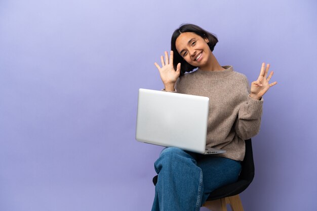 Giovane donna di razza mista seduta su una sedia con un computer portatile isolato su sfondo viola contando nove con le dita