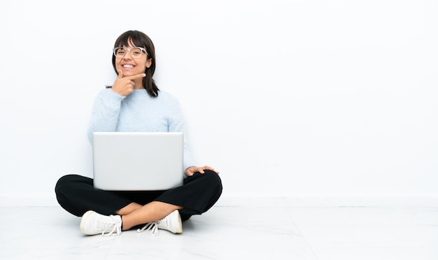 Giovane donna di razza mista seduta sul pavimento con il computer portatile isolato su sfondo bianco sorridente