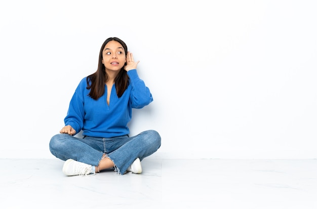 Giovane donna di razza mista seduta sul pavimento isolato su sfondo bianco ascoltando qualcosa mettendo la mano sull'orecchio
