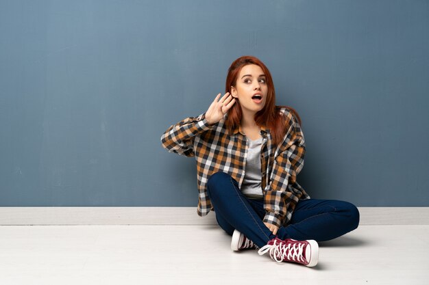 Giovane donna di redhead che si siede sul pavimento che ascolta qualcosa mettendo la mano sull'orecchio