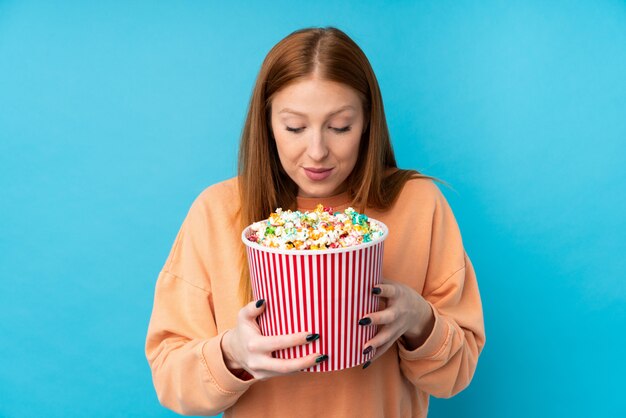 Giovane donna di redhead sopra la parete isolata che tiene un grande secchio di popcorn