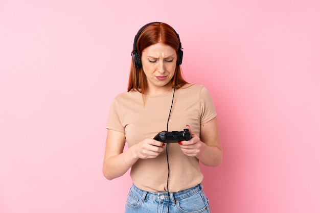 Giovane donna di redhead sopra la parete rosa isolata che gioca ai videogiochi