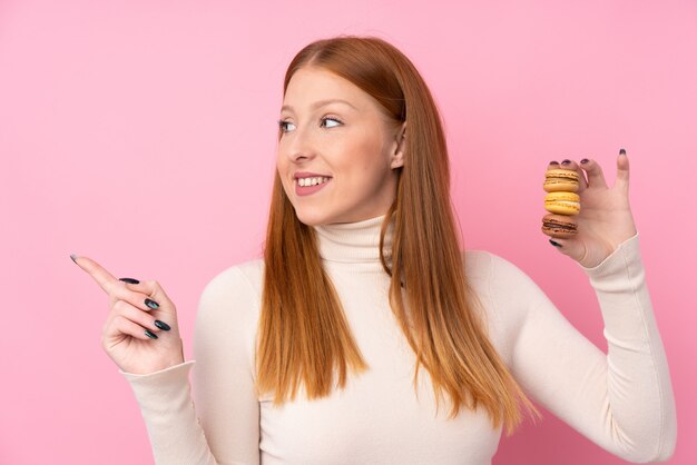 Giovane donna di redhead sopra la parete rosa isolata che tiene i macarons francesi variopinti e che indica lato