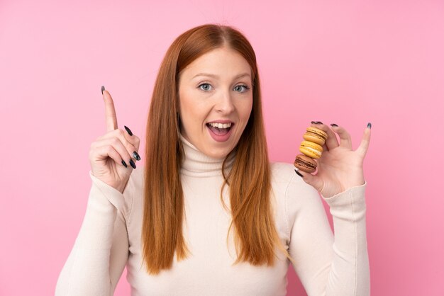 Giovane donna di redhead sopra la parete rosa isolata che tiene i macarons francesi variopinti e che indica su una grande idea