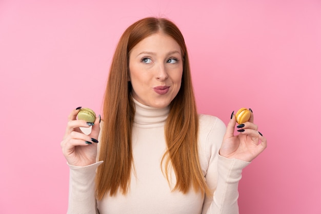 Giovane donna di redhead sopra la parete rosa isolata che tiene macarons e pensiero francesi variopinti