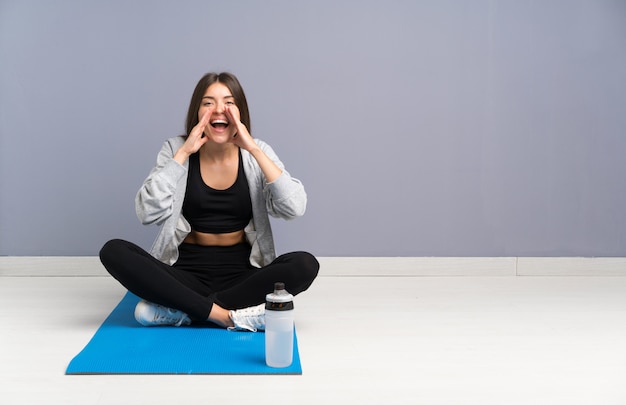 Giovane donna di sport che si siede sul pavimento con la stuoia che grida con la bocca spalancata