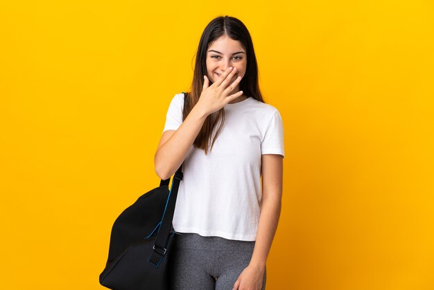 Giovane donna di sport con borsa sportiva isolata su giallo bocca di copertura felice e sorridente con la mano