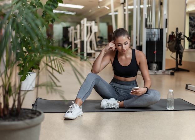 Giovane donna di sport in abiti sportivi facendo uso dello smartphone mentre sedendosi sulla stuoia nella palestra. Pausa allenamento. Concetto di stile di vita sano