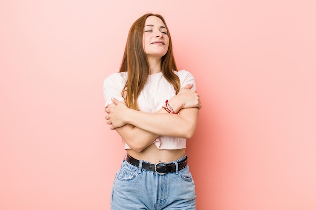 Giovane donna di zenzero rossa contro un abbracci di muro rosa, sorridente spensierato e felice.