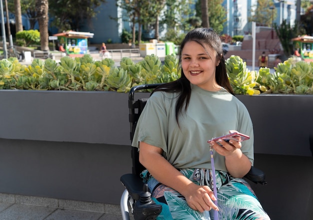 Giovane donna disabile positiva felice in sedia a rotelle che utilizza smartphone e sorride alla macchina fotografica mentre cammina all'aperto sulla soleggiata giornata estiva
