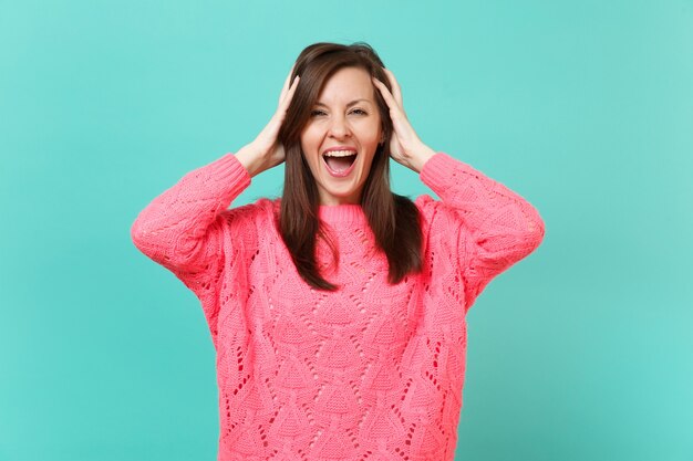 Giovane donna divertente allegra in maglione rosa lavorato a maglia che mette le mani sulla testa isolata sul fondo della parete turchese blu, ritratto in studio. Persone sincere emozioni, concetto di stile di vita. Mock up copia spazio.