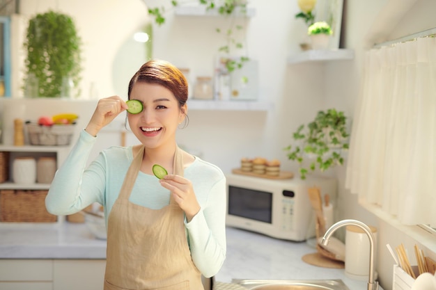 Giovane donna divertente con la fetta di cetriolo in cucina