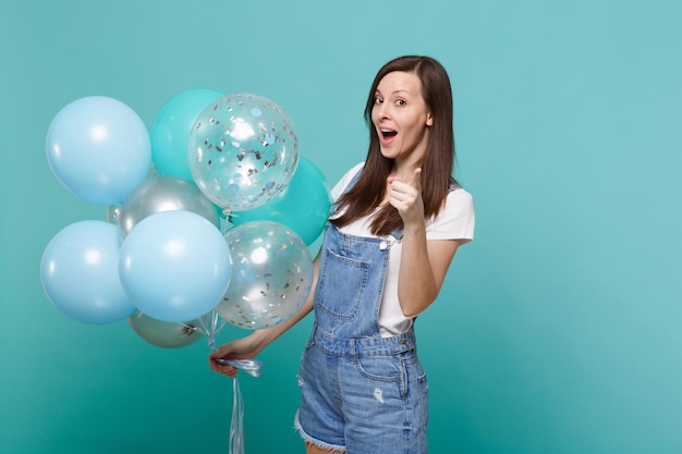 Giovane donna divertente in abiti di jeans che punta il dito indice sulla fotocamera, celebrando, tiene palloncini colorati isolati su sfondo blu turchese. Festa di compleanno, concetto di emozioni della gente.