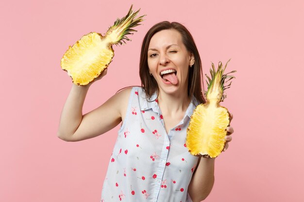 Giovane donna divertente in abiti estivi che mostra la lingua che tiene metà di frutta fresca di ananas maturo isolata su sfondo rosa pastello. Persone stile di vita vivido, relax concetto di vacanza. Mock up spazio di copia.