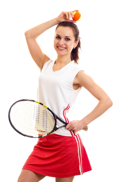 Giovane donna divertente in camicia sportiva bianca e gonna rossa con coda di cavallo castagna che tiene racchetta da tennis e palla gialla su sfondo bianco in studio