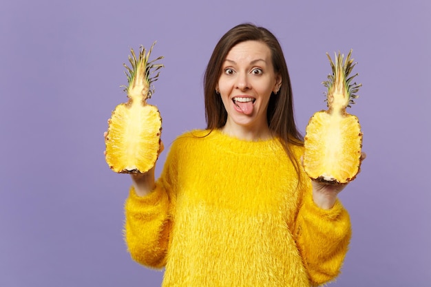 Giovane donna divertente in maglione di pelliccia che mostra la lingua che tiene due metà di frutta fresca di ananas maturo isolata su sfondo viola pastello. Lo stile di vita vivido delle persone si rilassa il concetto di vacanza. Mock up spazio di copia.