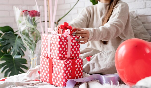 Giovane donna divertente seduta nel letto che celebra il giorno di San Valentino avvolgendo i regali