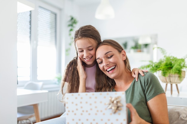 Giovane donna e ragazza a casa che celebrano la festa della mamma seduta sul divano figlia abbracciando madre baciare guancia mamma ridendo gioiosa azienda confezione regalo