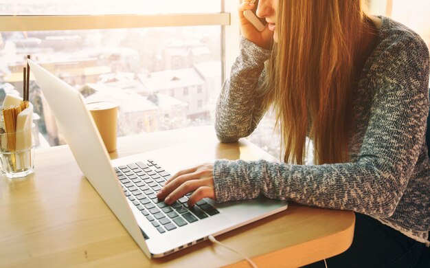 giovane donna è seduta al tavolo con un moderno computer portatile e parla al telefono