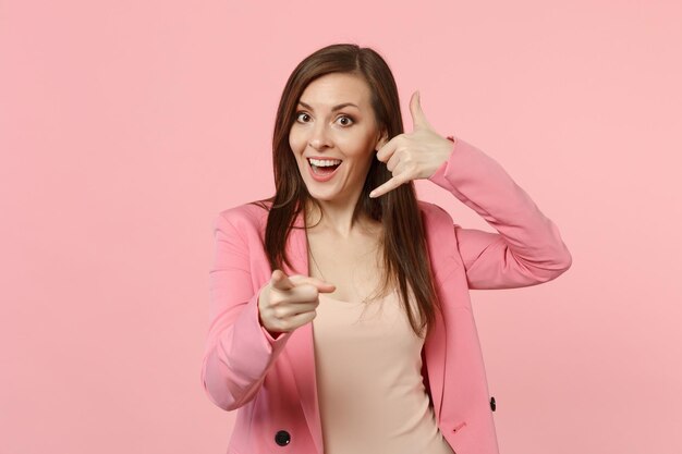 Giovane donna eccitata che fa un gesto telefonico come dice richiamami, puntando il dito indice sulla fotocamera isolata su uno sfondo rosa pastello. Persone sincere emozioni, concetto di stile di vita. Mock up spazio di copia.