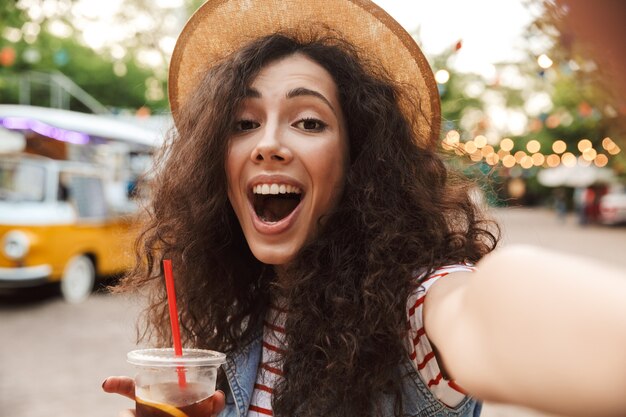 giovane donna eccitata con capelli castani ricci che indossa un cappello di paglia estivo sorridente e bere tè freddo da un bicchiere di plastica all'aperto, mentre scatta foto selfie