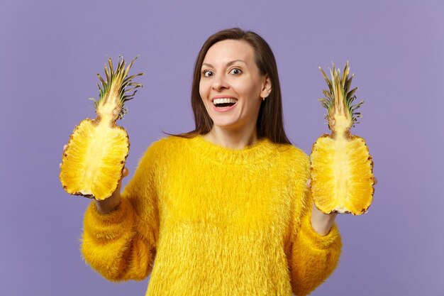 Giovane donna eccitata in maglione di pelliccia che tiene in mano metà di frutta fresca di ananas maturo isolata su sfondo viola pastello. Persone stile di vita vivido, relax concetto di vacanza. Mock up spazio di copia.