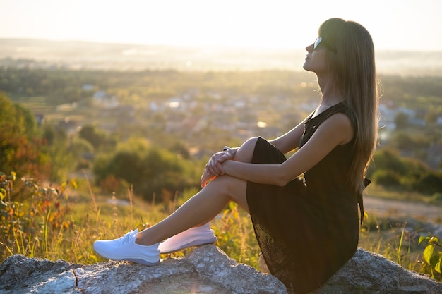 Giovane donna elegante in abito corto nero e scarpe da ginnastica bianche seduta su una roccia che si rilassa all'aperto la sera d'estate. Signora alla moda che gode del caldo tramonto in natura.