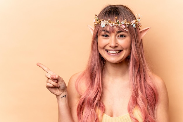 Giovane donna elfo con capelli rosa isolati su sfondo beige sorridente e indicando da parte mostrando qualcosa in uno spazio vuoto