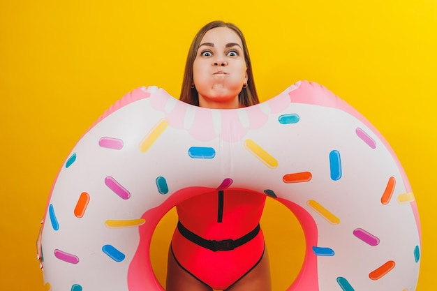 Giovane donna emotiva che indossa un costume da bagno rosa con un anello gonfiabile isolato su uno sfondo giallo brillante Estate hotel piscina mare vacanza sole concetto di abbronzatura