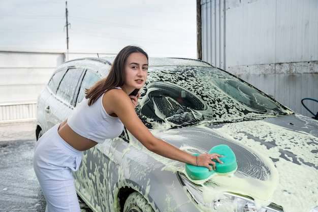 Giovane donna esile che pulisce la sua auto utilizzando una schiuma ad alta pressione e una spugna per autolavaggio.