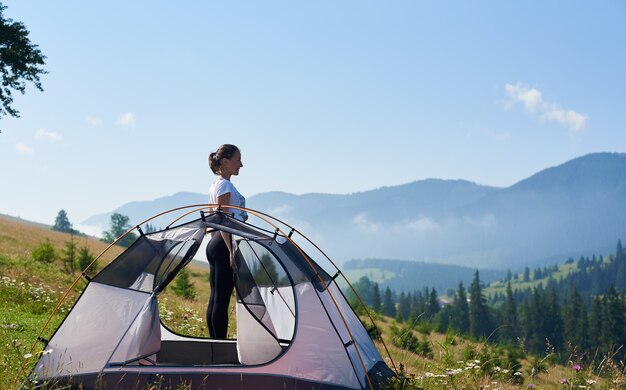 Giovane donna esile della viandante che sta alla piccola tenda turistica che gode di bello panorama delle montagne coperte di nuvole bianche sotto il chiaro cielo blu sulla mattina luminosa dell'estate. Turismo e concetto di campeggio.