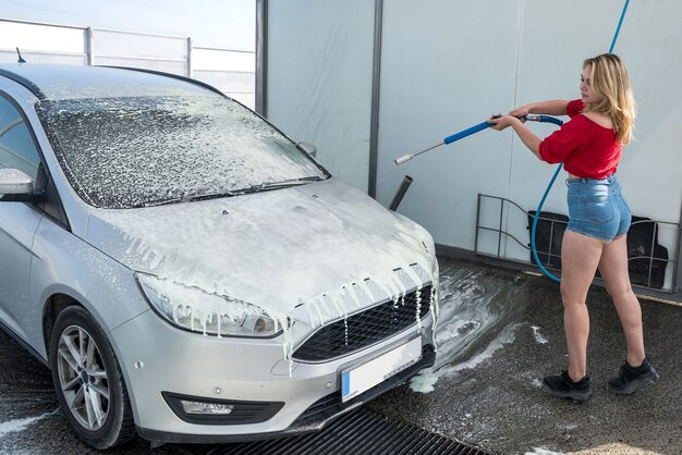 Giovane donna esile sessuale che utilizza tubo flessibile con schiuma spray per pulire la sua auto dallo sporco