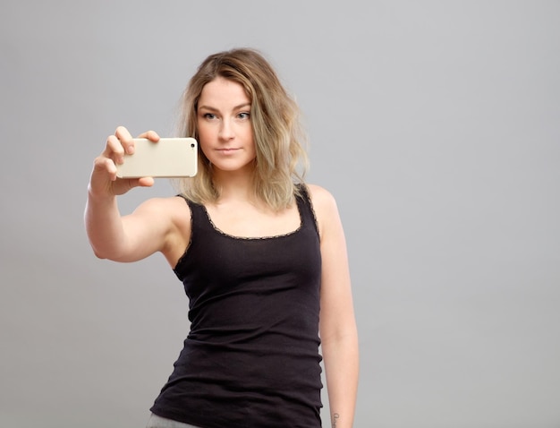 Giovane donna espressiva scattare foto tramite cellulare su sfondo grigio