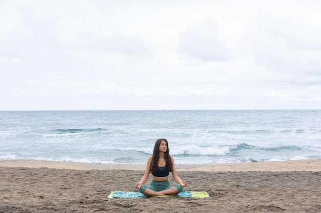 Giovane donna etnica seduta in riva al mare nella posa di Padmasana durante la pratica dello yoga