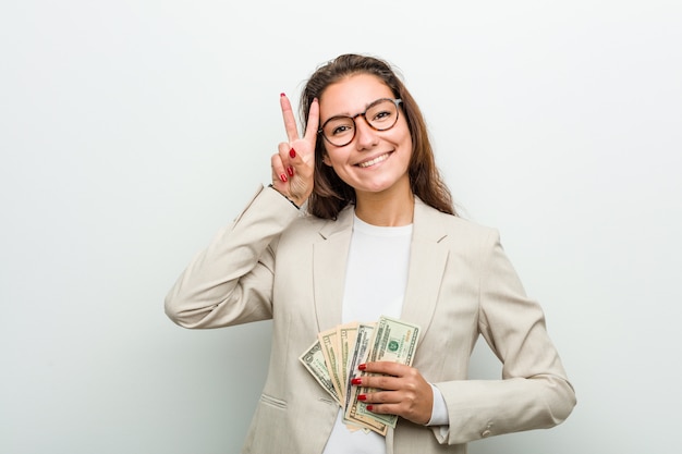 Giovane donna europea di affari che tiene le banconote del dollaro che mostrano il segno di vittoria e che sorridono ampiamente.